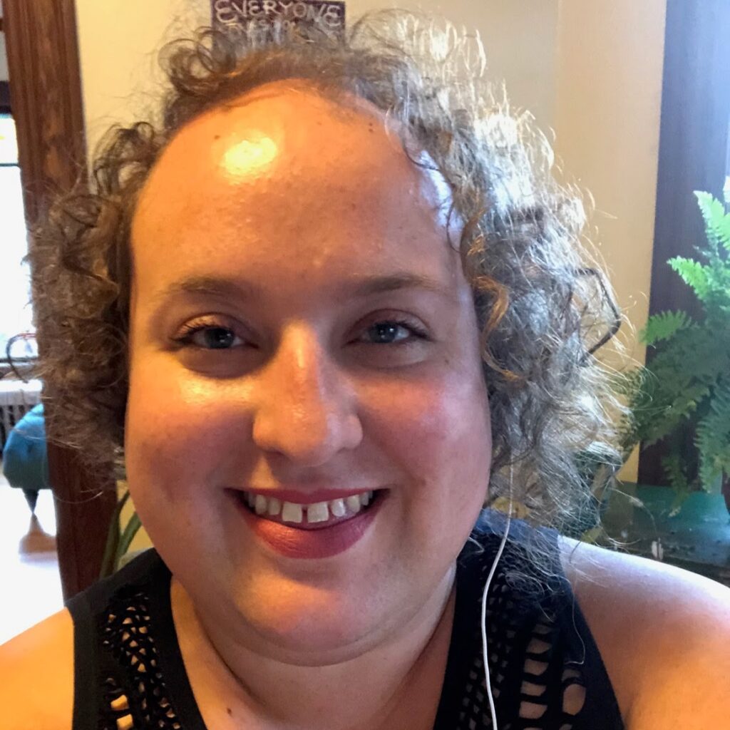 Image shows a white woman with curly gray hair smiling and wearing a black tank top. There is a doorway and plant in the background.