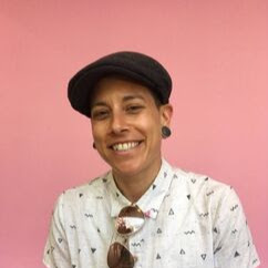 Image shows a white person smiling and wearing a black newsboy cap and a white patterned collared shirt with sunglasses hanging on the collar. They have black gauge earrings and are standing in front of a pink background.