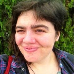 Image shows white woman with short brown hair smiling wearing a purple and black flannel shirt.