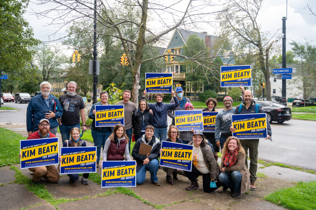 Image shows a large group of white people of all ages, standing outside in a neighborhood holding Kim Beaty yard signs. 