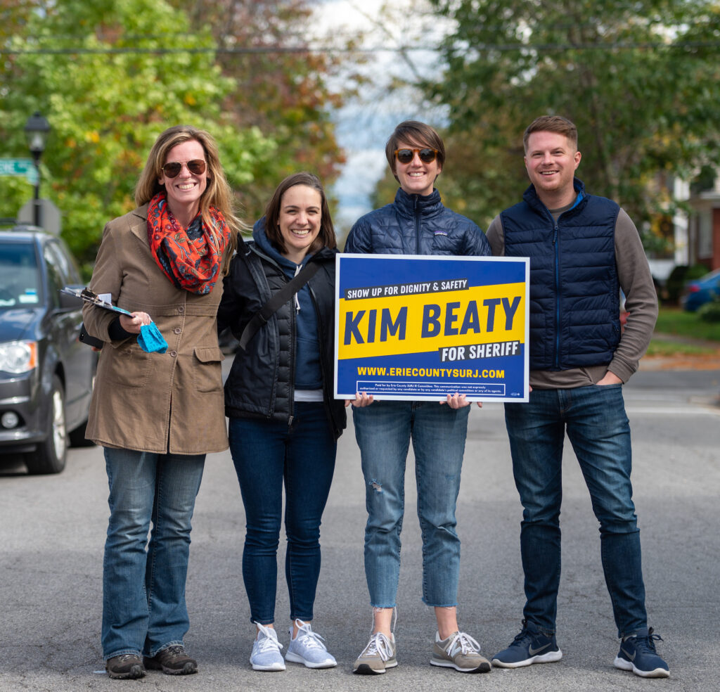 Image shows a group of 4 white people who look to be in their 30's, standing outside. One of them is holding a Kim Beaty yard sign.