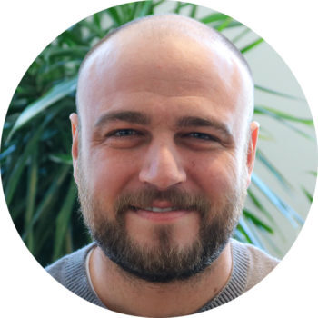 Image shows a white man with a shaved a head and brown short beard smiling in front of a plant.