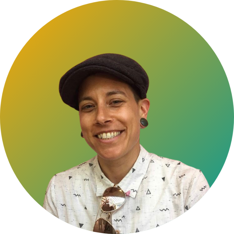 Image shows a white nonbinary person smiling in front of a yellow-green gradient background. They are wearing a white button up shirt with a small black print, plugs in their ears, sunglasses hanging from their shirt and a black newsboy cap.