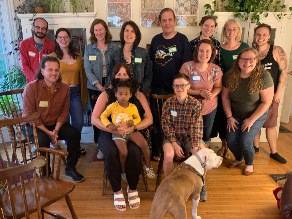 photo shows a group of 13 white folks posing for a group photo inside a home on a wood floor