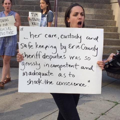 Image shows a white woman at a rally, holding a sign and shouting. The sign reads "...her care, custody and safe keeping by Erie County sheriff deputies was so grossly incompetent and inadequate as to shock the conscience." 