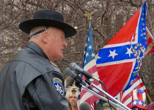 Image shows an older white man wearing a sheriff's uniform, speaking at a public event. There are American flags and confederate flags in the background.