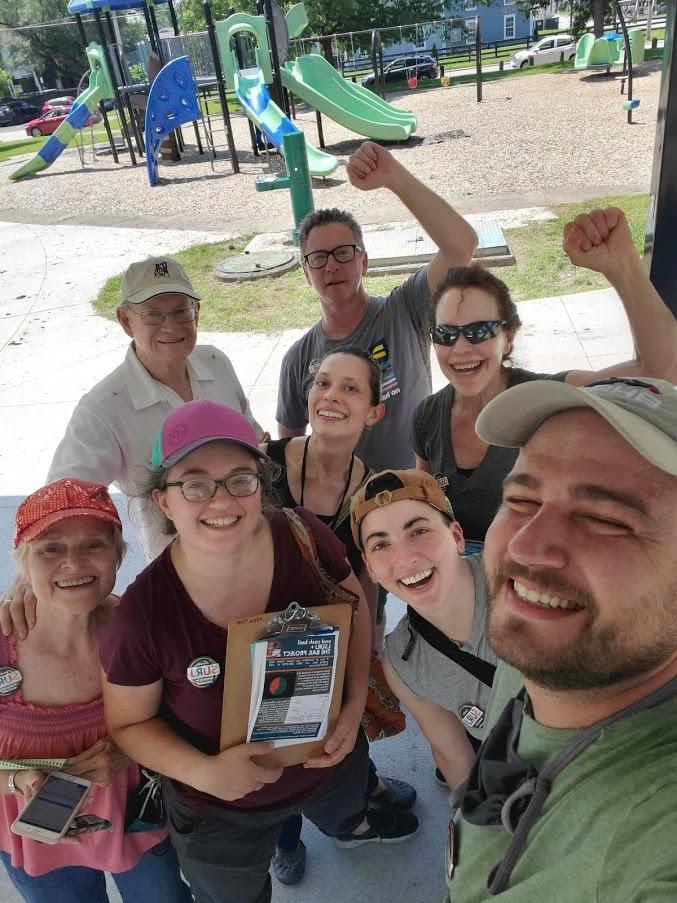 image shows a group of white folks in a park posing for a selfie. One of them is holding a clipboard with canvassing literature.