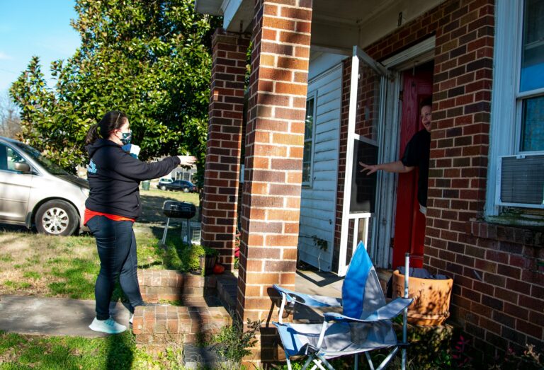 image shows a white woman wearing a mask speaking with another white woman at her front door