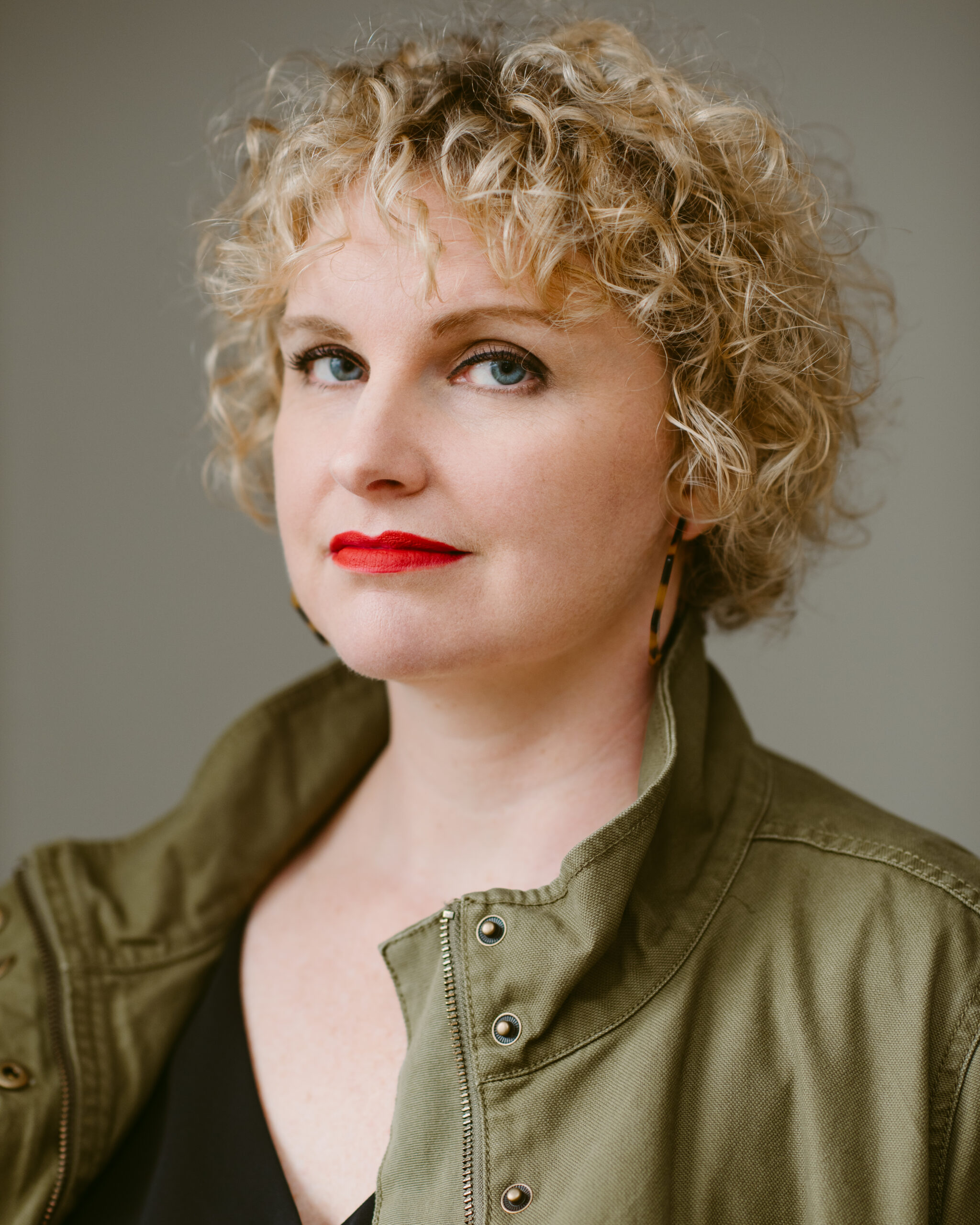 image shows a white woman with short blonde curly hair. She's wearing red lipstick and a green jacket.