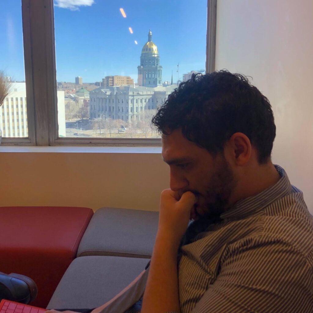 photo shows a brown man with a beard holding his hand to his face, looking away from the camera. Out of the window is a state capitol building.