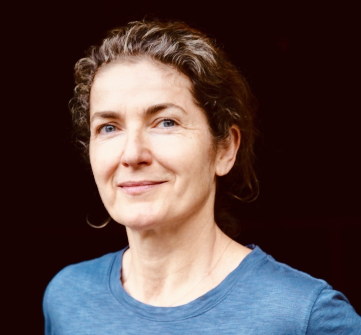 Photo shows a white woman wearing a blue shirt, with a black background. She is smiling slightly and has short brown hair.