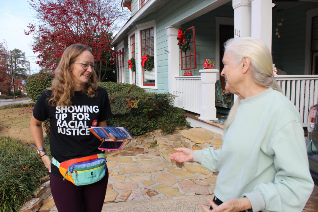 Julia, a white woman with long blonde hair wears a black Showing Up for Racial Justice shirt. She talks to an older woman with white hair and a light green sweatshirt in front of a light blue single-family home.