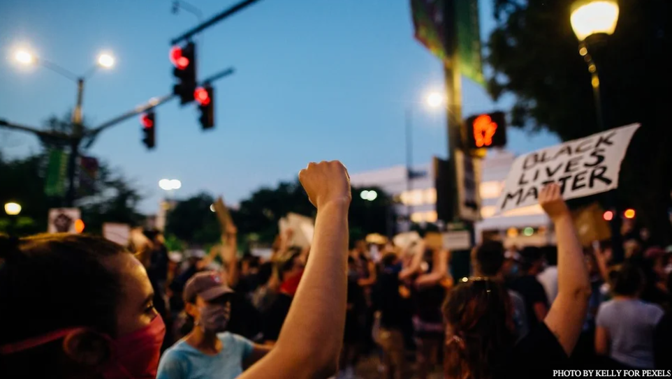 Image of a white person's arm raised in a fist at a protest outside under a blue sky and streetlights, surrounded by people.