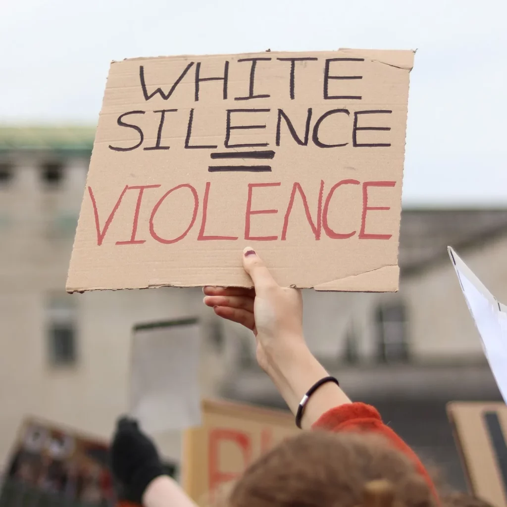 Image of a white person's hand holding up a cardboard sign with "white silence = violence" written in red and black marker.