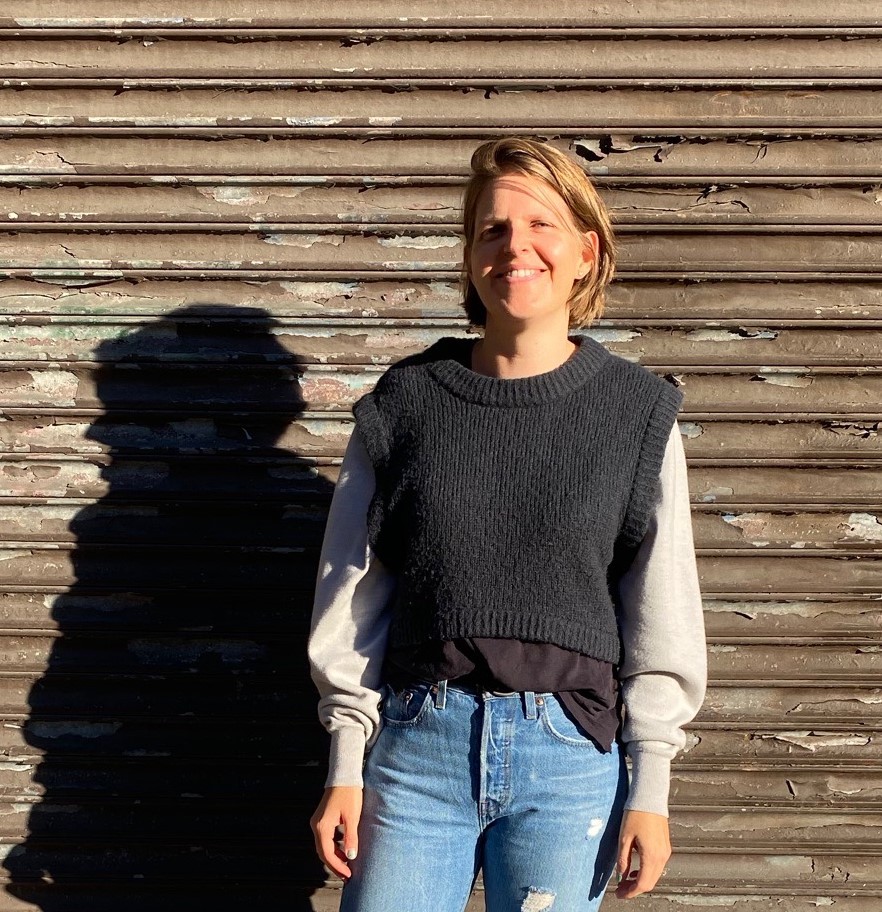 Image shows white woman with short blonde hair smiling and wearing a black sweater with white sleeves and jeans in front of metal wall.