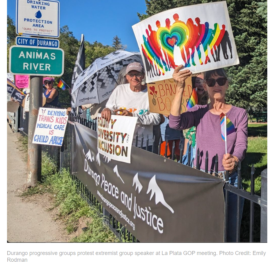 Image of white protesters standing on grass in front of a blue sky holding pro-LGBT, pro-democracy signs.