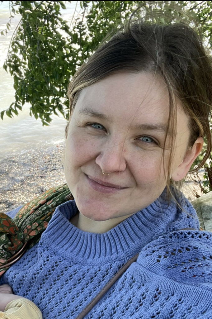 Image shows white woman with long dark blonde hair tied back wearing a purple sweater smiling with a tree in the background.