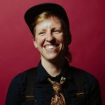 Image shows a white person with short blonde hair wearing a black baseball hat, black collared shirt and a patterned tie. The person is smiling and standing in front of a red wall.