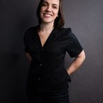 Image shows a white woman with short brown hair smiling and wearing a short sleeved black dress standing in front of a gray background.