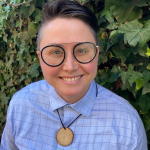 Image shows a masc white person wearing a blue collared shirt, glasses with round black frames, standing in front of a hedge. They are smiling.