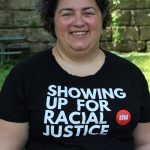 Image shows a white woman wearing a black tshirt that says "showing up for racial justice." She is outside, is smiling, and has short dark curly hair.