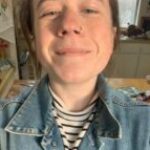 Image shows white woman with brown hair tied back smiling and wearing a denim jacket and a black and white striped shirt.