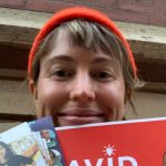 image shows a white woman smiling, wearing an orange beanie. She is holidng campaign literature in front of her face.