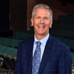 Picture shows a white man with short gray hair wearing a blue suit and tie.