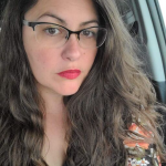 Image shows a white woman with long brown wavy hair wearing glasses and a floral blouse.