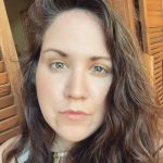 Photo shows picture of a white woman with long brown wavy hair wearing a white shirt standing in front of wood paneling.